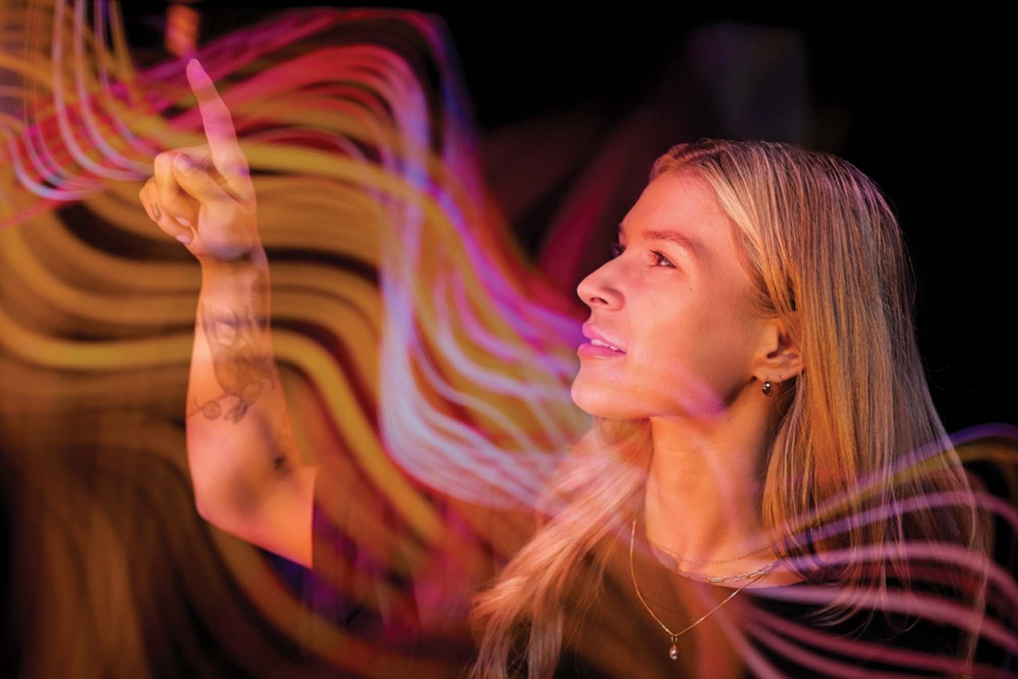 A woman is captured in a moment of wonder, her profile illuminated by the vibrant light trails swirling around her hand.