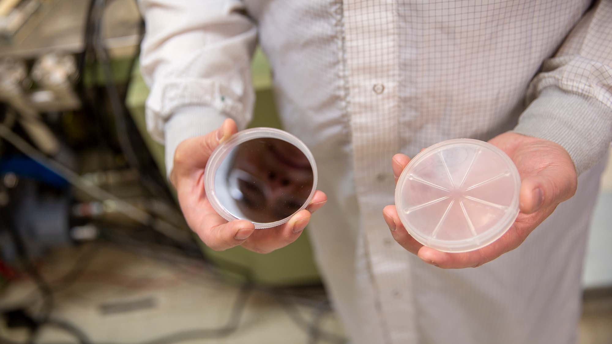 A person in a white lab coat holds two petri dishes towards the camera; one dish is covered with a reflective film and the other is segmented into sections.
