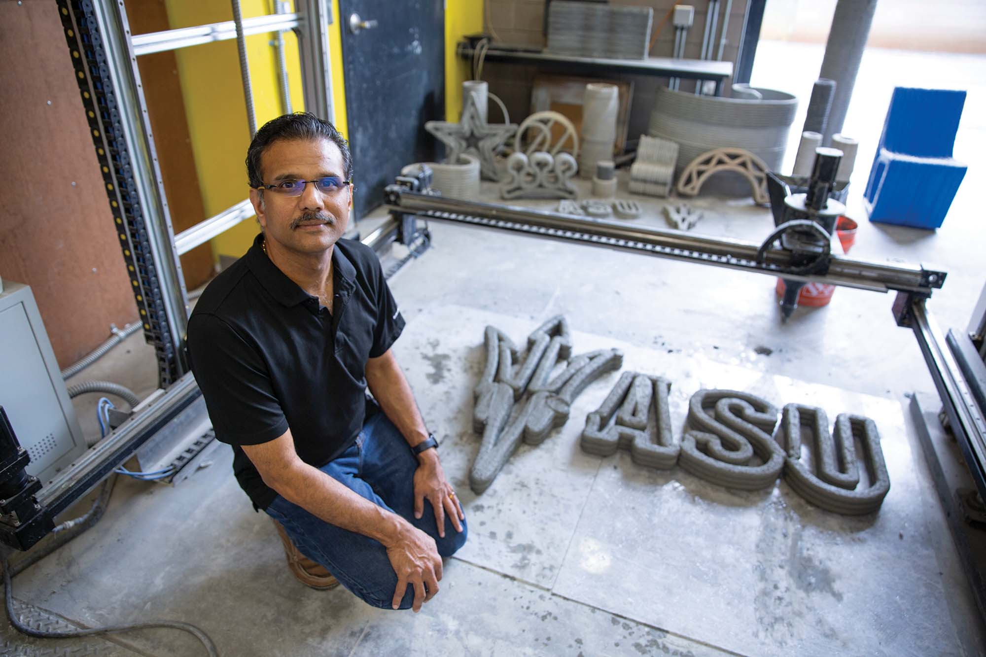 Fulton Professor of Structural Materials Narayanan Neithalath, wearing a black polo shirt and jeans, kneels next to a large, textured 3D print of the letters "ASU" with the pitchfork logo in a workshop.