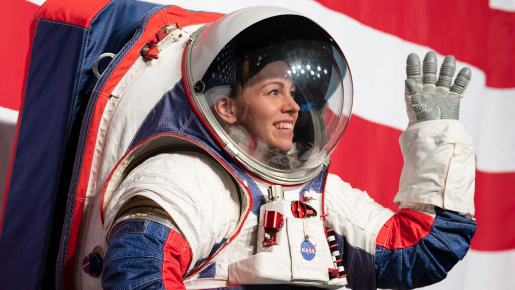 A woman stands inside a modern red, white, and blue NASA spacesuit, smiling broadly and waving, in front of the red and white stripes of the American flag.