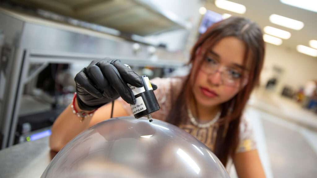 A woman, Ramos, holds a stubby, cylindrical instrument against a dome of glass coated with Swift Coat's coating, taking a measurement, in a lab.