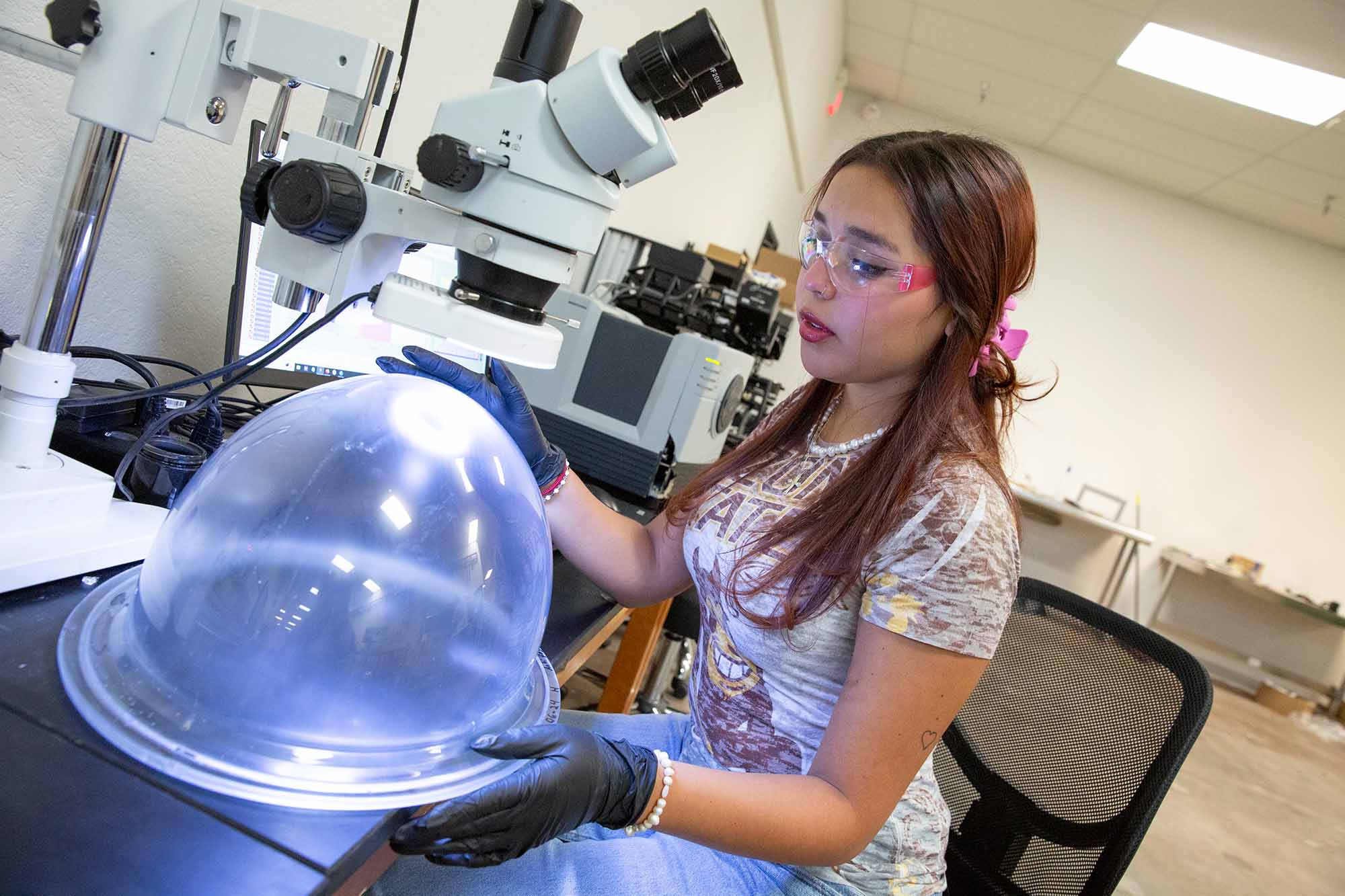 An ASU student wearing safety goggles and gloves is working with a large, clear dome under a microscope in a laboratory.