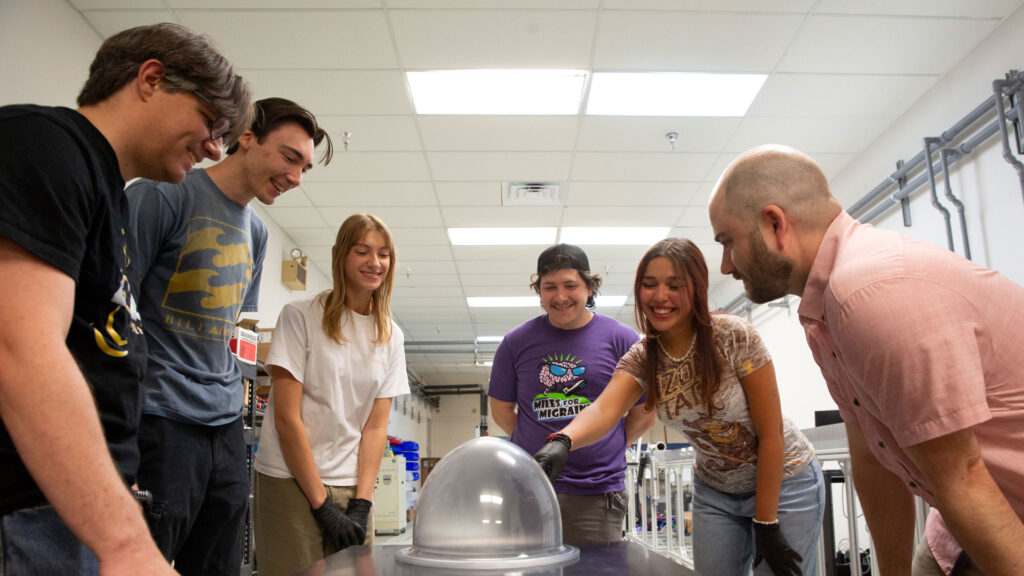 A group of six people stand together around a glass dome coated in Swift Coat's coating while one person, Abril Ramos, points to it. They are in a lab.