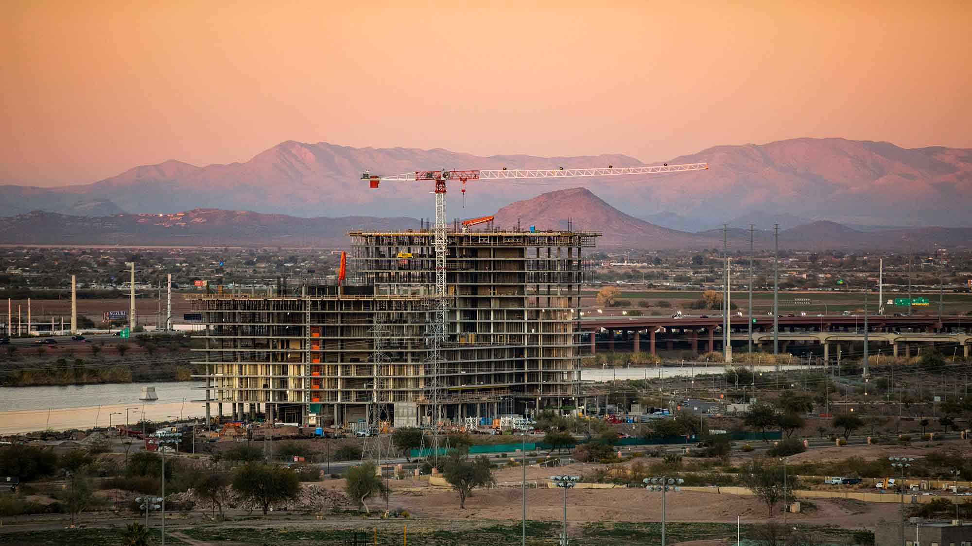 This image captures a construction site at sunset, with a tall building framework and a crane towering above it.