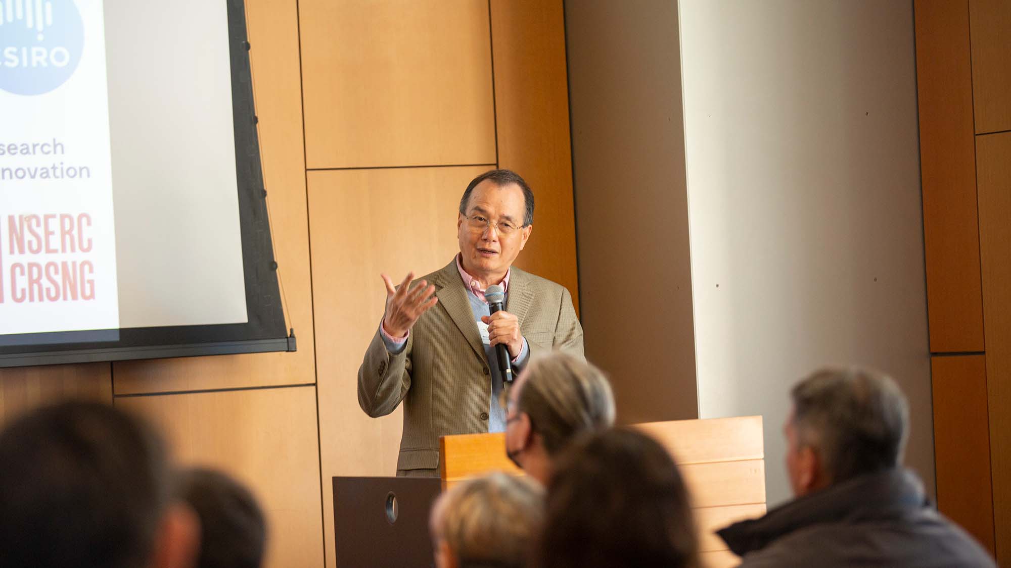 This image features a man speaking at a podium, holding a microphone and gesturing with his hand as he addresses an audience.