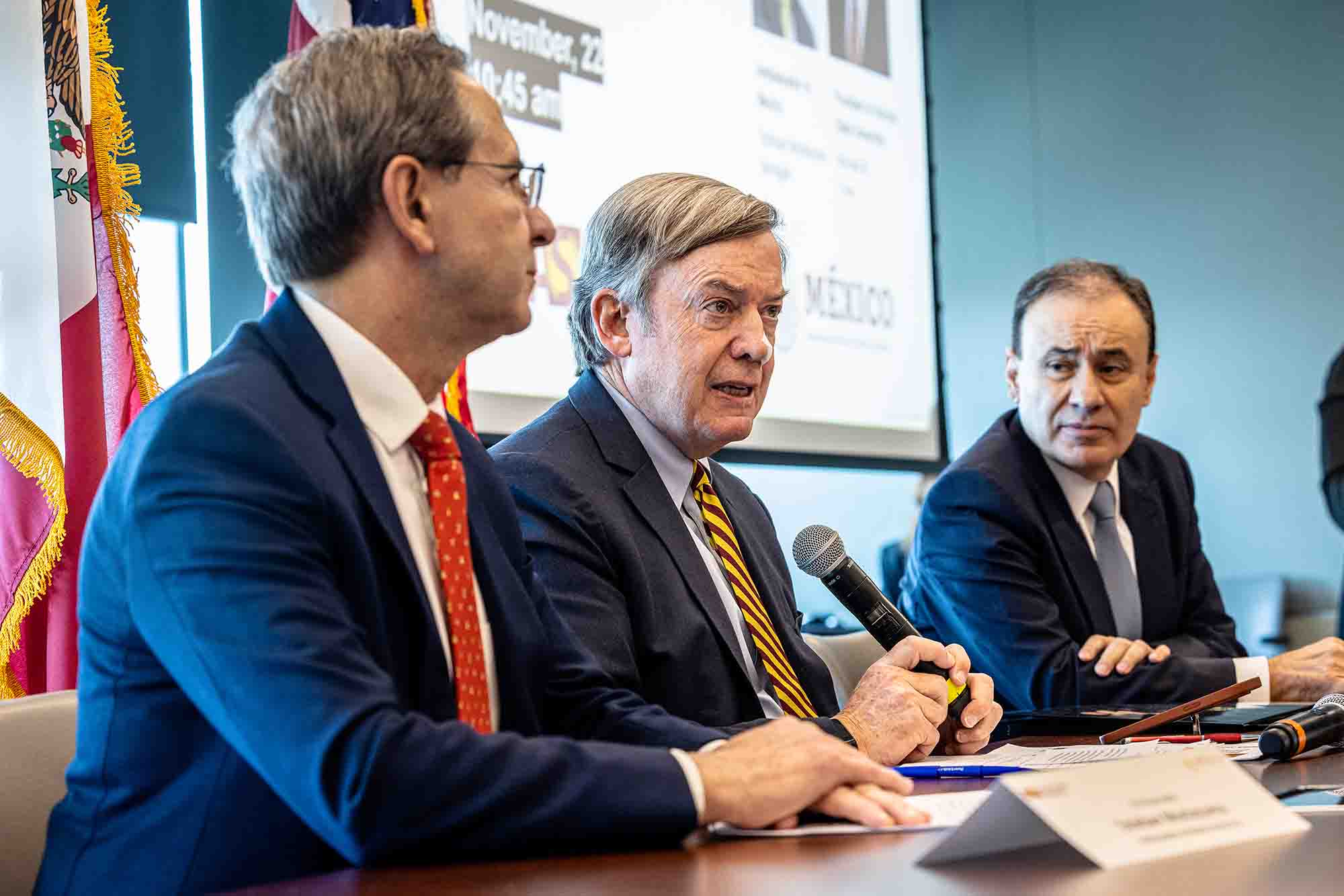 The image shows three men in formal business attire seated at a conference table, engaged in a discussion.