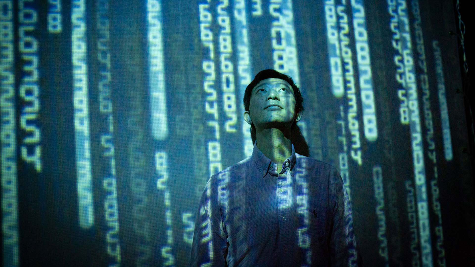 A man looking upward thoughtfully with glowing blue lines of binary code projected on him and the background.