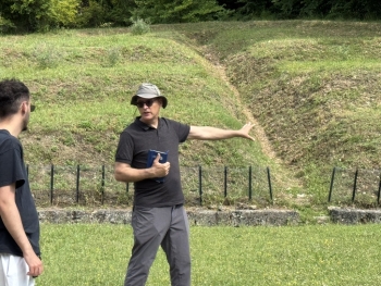 A person wearing a hat gestures toward a grassy slope while speaking to another person outdoors.