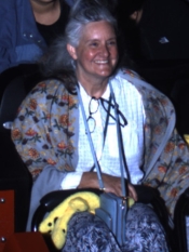 A smiling woman with long gray hair sits indoors wearing a patterned shawl and holding a small bag.