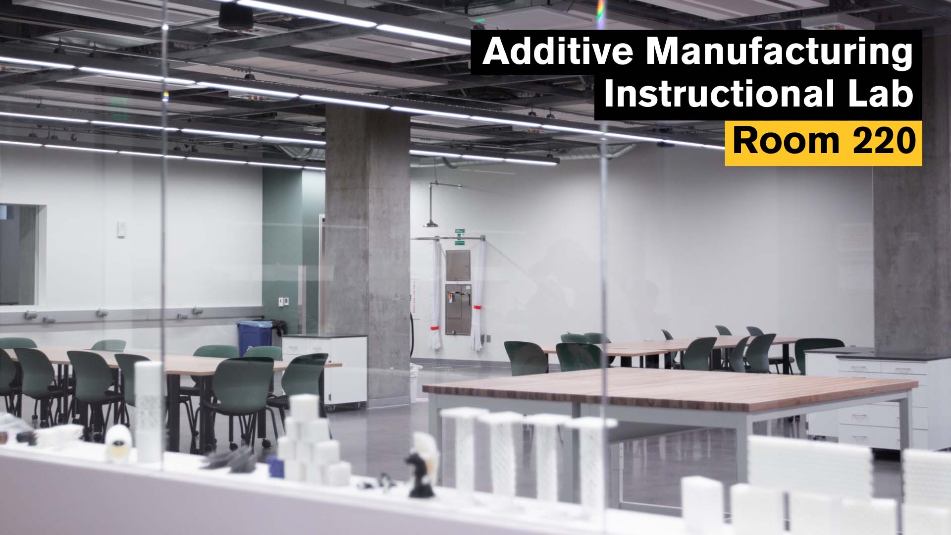 Additive Manufacturing Instructional Lab, Room 220. A modern instructional lab space with large worktables, green chairs and exposed industrial ceilings. 3D printed parts are displayed on the window sill, and the room is set up for collaborative teaching and hands-on prototyping.