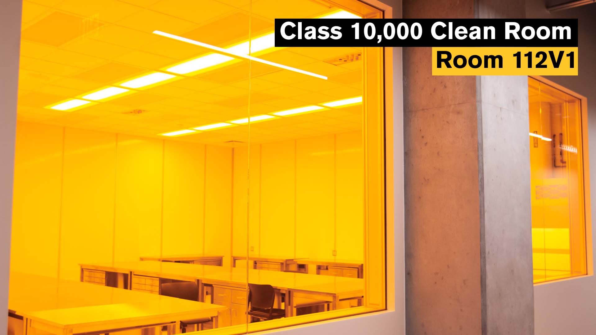 Class 10,000 Clean Room, Room 112V1. A cleanroom environment with bright yellow-orange filtered lighting, empty stainless steel worktables and ergonomic chairs. Large viewing windows and a sealed environment highlight contamination-controlled workspace.