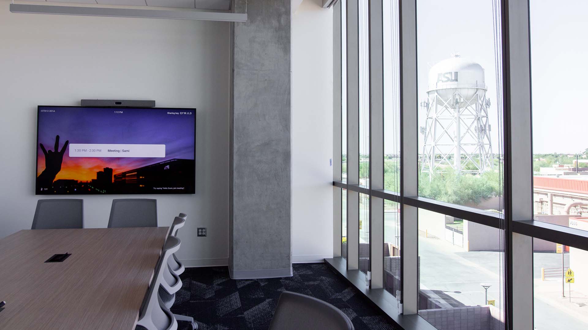 A modern conference room with a large window wall overlooking an outdoor scene. The windows are tall and paneled with a view of an ASU water tower and a flat desert landscape outside. Inside, a light wooden conference table is partially visible in the foreground, with several gray chairs around it. A large, wall-mounted display screen shows a meeting interface with a sunset background.