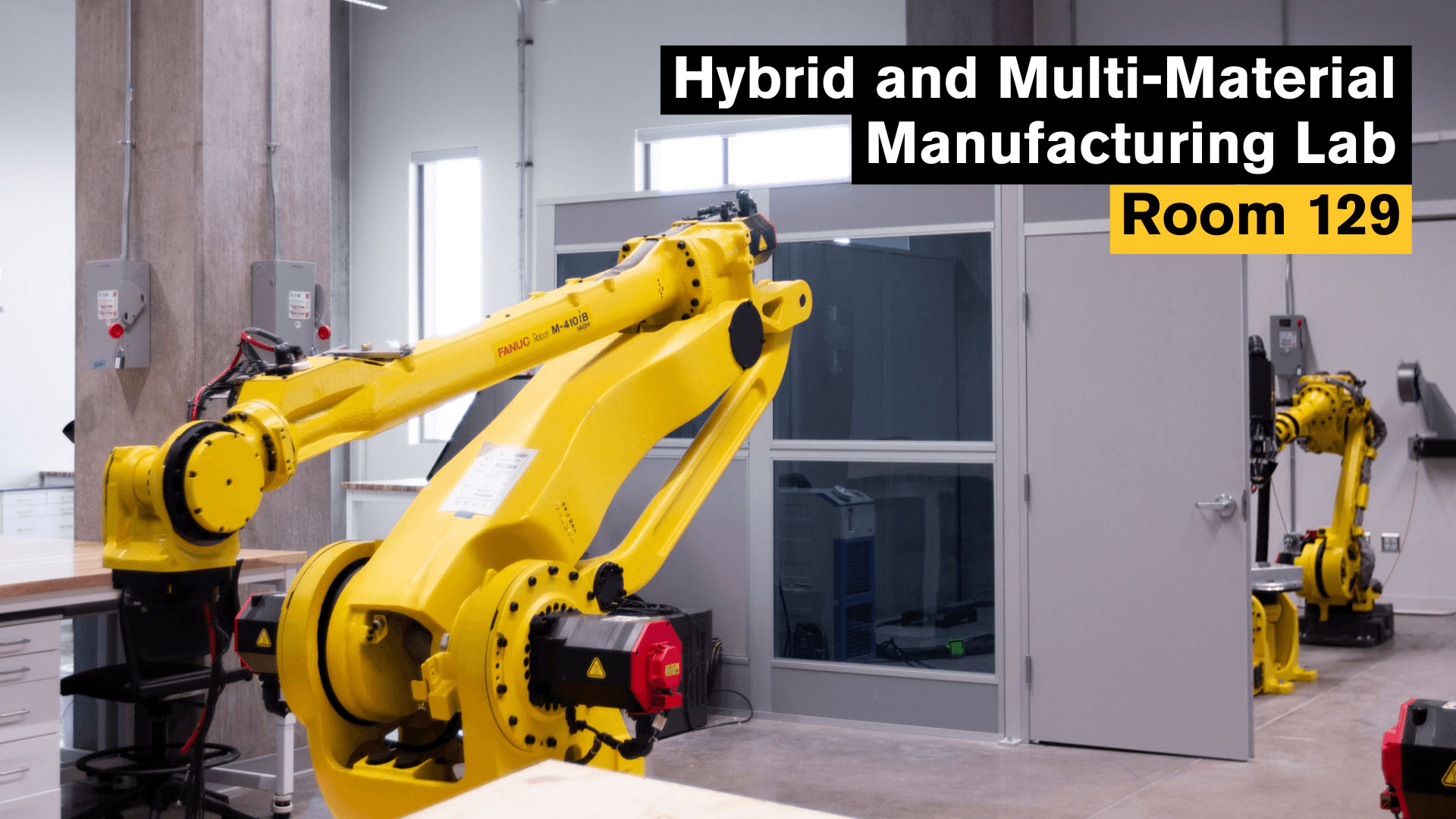 Hybrid and Multi-Material Manufacturing Lab, Room 129. A robotics lab with large FANUC yellow robotic arms, placed near worktables and a partitioned office space. The environment is designed for testing hybrid and multi-material manufacturing systems.